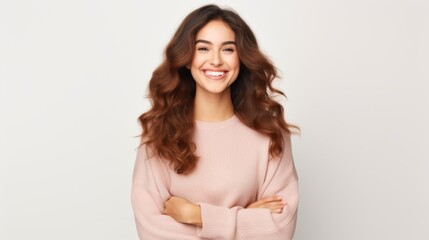 Obraz premium A young woman with long, wavy hair and a soft, appreciative smile, wearing a light pink sweater, stands confidently with her arms crossed against a light background.