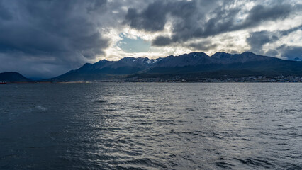 A picturesque snow-covered mountain range of the Andes against the sky and dark clouds. The city buildings of Ushuaia are at the foot. In the foreground is the surface of the ocean. The Beagle Channel