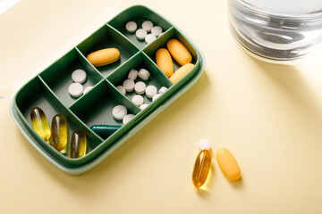 Pill box with various pills, vitamins and dietary supplement next to water glass and omega 3 capsule.