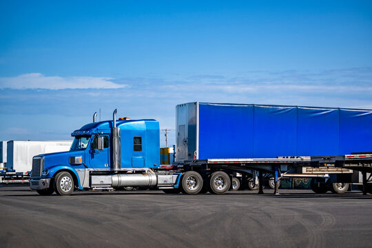 Bright blue big rig bonnet semi truck tractor with conestoga semi trailer standing on the warehouse parking lot waiting for the next loads