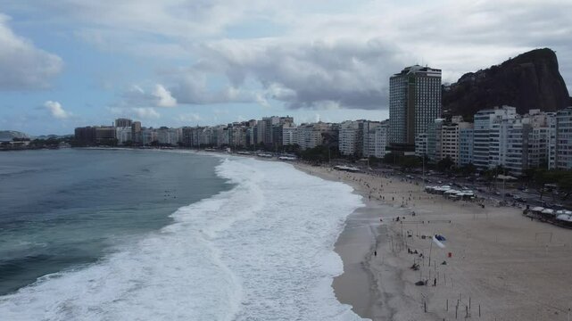 V&iacute;deo a&eacute;reo da praia de Copacabana, Rio de Janeiro, em um dia nublado, destacando o cal&ccedil;ad&atilde;o ondulado, o mar tranquilo, a areia dourada e o contraste entre a cidade e o clima cinzento