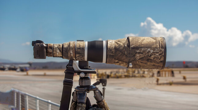 A camera with a large telephoto lens wrapped in camouflage on a tripod at an airshow