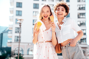 Two young beautiful smiling hipster female in trendy summer clothes. Carefree women posing on street background. Positive models eating tasty ice cream in waffles cone in sunny day, cheerful and happy