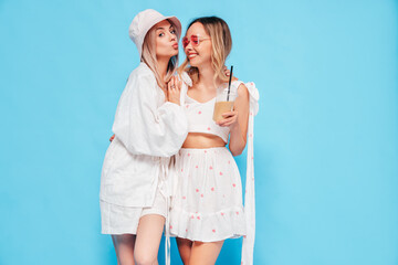 Two young beautiful smiling blond hipster female in trendy summer clothes. Carefree women posing near blue wall in studio. Positive models having fun. Cheerful and happy, sunglasses