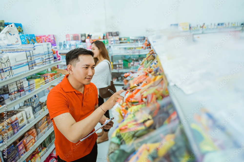 Wall mural male shop assistant records items with a list on a clipboard while working at minimarket