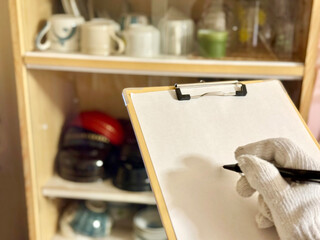 お椀の入った食器棚の前でクリップボードに記入する軍手をはめた手