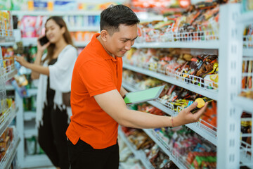 male waiter holding product on shelf while checking with carrying computer tablet in supermarket