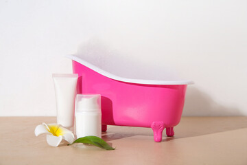Small bathtub with foam and cosmetic products on color table against light background