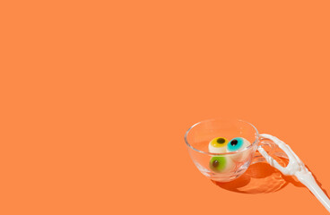 Skeleton hand is holding glass with eyeballs, orange background. Minimal halloween food or drink concept.