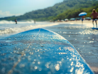 A close-up view of a blue surfboard resting on wet sand, with sparkling water droplets reflecting sunlight. In the background, surfers are seen riding waves and beachgoers are enjoying the sunny day.