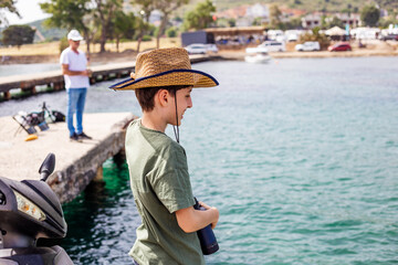Teenager boy in a straw hat, holding binoculars, smiling by the sea
