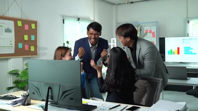 Diverse team of business professionals is cheering and clapping, gathered around a computer in their modern office. Their enthusiastic celebration suggests a significant achievement