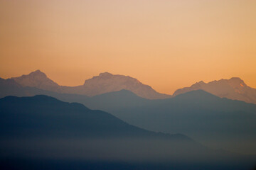 A serene dawn breaks over the horizon, casting a soft golden hue across the majestic snow-capped peaks. The layered silhouette of the mountains fades into the distance, their gentle ridges cloaked in 
