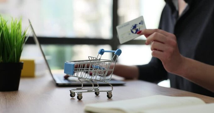 Woman enters card details to make purchase sitting at table with toy shopping cart. Concept of modern e-payment through online system slow motion