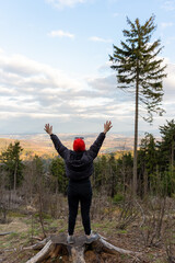 Woman in red beanie with open arms enjoying a panoramic mountain view from a forested hilltop on a clear day. Freedom, adventure, and connecting with nature during a scenic outdoor exploration. . High