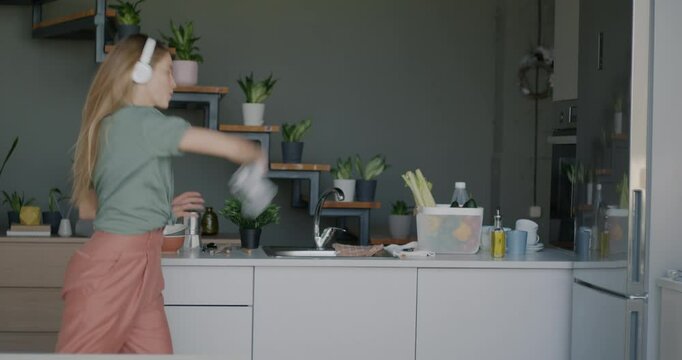 Slow motion of playful young lady having fun dancing listening to music with headphones in kitchen in modern house. Youth culture and leisure activity concept.