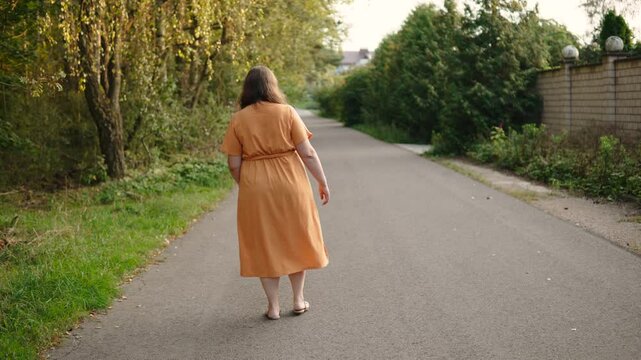 Lonely woman limping walks along the road, back view. Female person injured her leg and goes home outdoors. Domestic trauma.