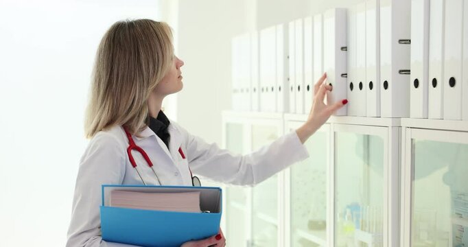 Focused female doctor looks at ring binders for necessary document case with data about patient. Organized folder structure and archive on light rack slow motion