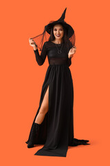 Beautiful young happy Asian woman dressed as witch for Halloween on orange background