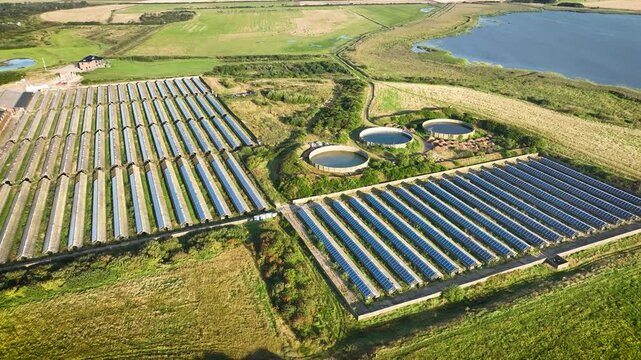 Aerial footage of sprawling solar farm with rows of solar panels installed on roofs of animal breeding farm along with three slurry storage facilities, contributing to sustainable energy generation.