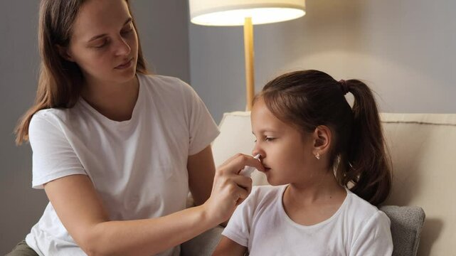 Caring mother using nasal spray to treat her little girl&rsquo;s congestion while daughter with stuffy nose resting on the sofa feeling sick catching cold having flu symptoms at home