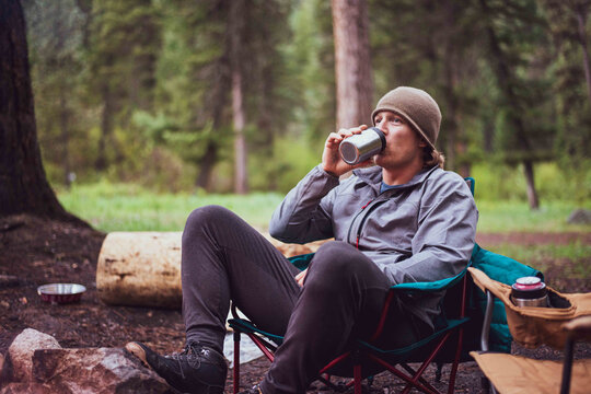Man sitting in a camp chair around a fire drinking from a can