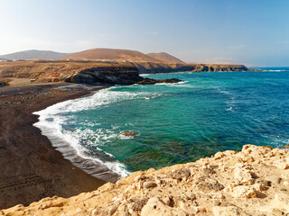 A beach with black volcanic sand in the town of Ajuy on the island of Fuerteventura is shown in this image. The rugged coastline meets clear turquoise waters, while barren hills rise in the background