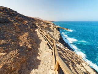 A walking path in the town of Ajuy, on the island of Fuerteventura, runs along a rugged cliffside. It overlooks the deep blue ocean, with waves crashing against the rocks and the horizon stretching fa