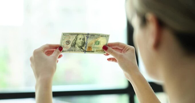 Woman with red manicure checks authenticity of dollar banknote holding bill against sunny light from window. Fake money concept. Criminal liability
