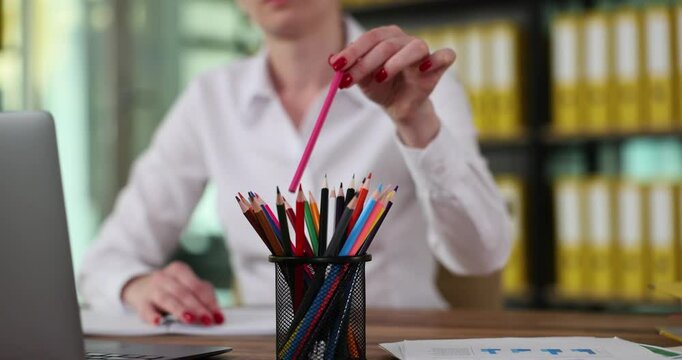 Female manager smoothly takes pink pencil out of holder with colored pencils. Senior worker makes notes on paper about mistakes in subordinate report