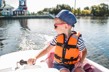 A handsome, fashionable little boy of 5 years old in a bright life jacket and sunglasses drives a...