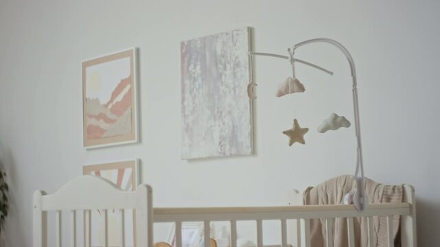 No people shot of cozy baby bedroom interior in pastel colours with interactive soft toys mobile hanging above white wooden crib and pictures on wall
