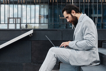 Man with beard wearing gray suit, working on laptop while sitting on outdoor steps. Reflected building glass and modern architecture in background is visible