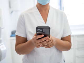 An unrecognizable healthcare professional in a white uniform and mask checks the smartphone while...