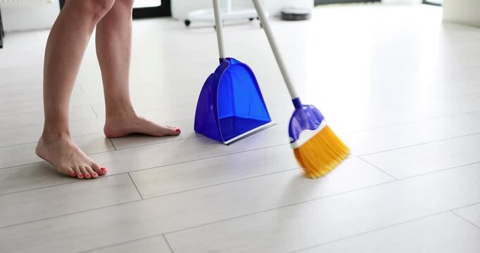 Barefoot female person sweeps light wooden floor using broom and dustpan. Housewifely woman cleans up bright room. Household chores. Every day cleaning routine