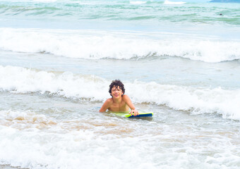 Brunette teenager male surfing on the waves of the South China Sea. Young kid on surf board laughing. Active water sports for teenagers. Surfer boy swimming with body board