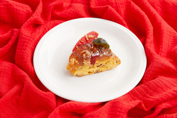 A slice of festive cake with candied fruit and nuts on a white plate, set against a red fabric backdrop