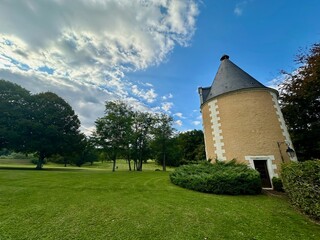 Historic stone tower in the middle of a large, lush green park on a sunny day with a bright blue sky and scattered clouds, surrounded by trees and well-manicured lawns in peaceful countryside setting