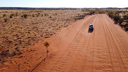 Aerial picture of a car driving in the middle of the outback. Long sandy road with bushes on the side. 4wd adventure in the red centre of Australia. Road sign on the side of the road.