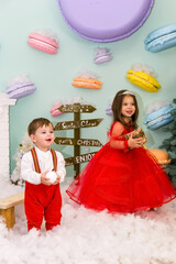 A little girl and a boy in red clothes have fun in the artificial snow by the fireplace in the studio. The girl is holding a box with gifts. Little kids in red christmas costume having fun beside a