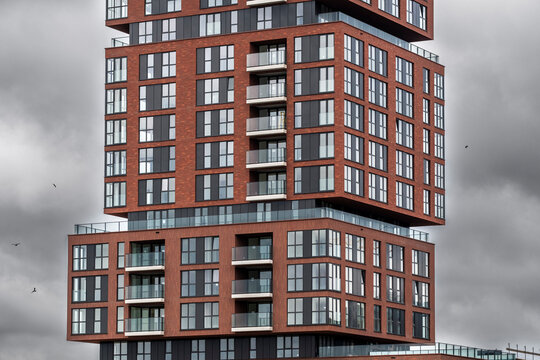 A striking building made of red bricks is depicted with its staggered and stacked design, showcasing modern architecture with numerous balconies under a cloudy sky.