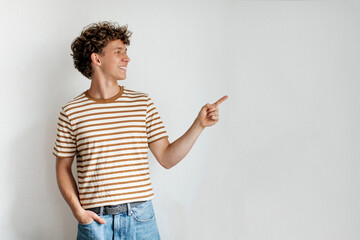 A young man with curly hair stands casually, wearing a striped shirt and jeans. He smiles and...