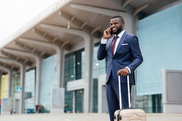A well-dressed businessman stands in front of an airport terminal, smiling while talking on his...