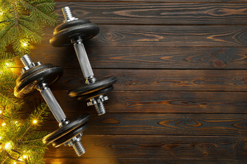 Fototapeta premium Dumbbells, fir tree branches and Christmas lights on wooden table, flat lay. Space for text