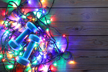 Dumbbells, and Christmas lights on wooden table, top view. Space for text