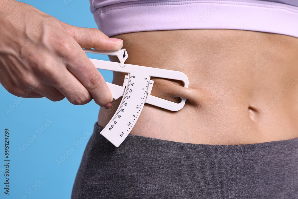 Wall mural Woman measuring body fat with caliper on light blue background, closeup