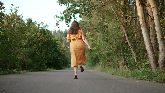 Plus size woman happily bouncing on leg on road in suburbs, back shot. Happy female person in orange dress with phone in hand is happy. Concept of body positivity, self-acceptance.