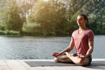 Fototapeta premium A man sits cross-legged on a yoga mat near a tranquil lake, enjoying a moment of meditation while surrounded by lush greenery under bright sunlight.