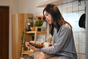 Peaceful young asian woman sitting at kitchen table and reading book