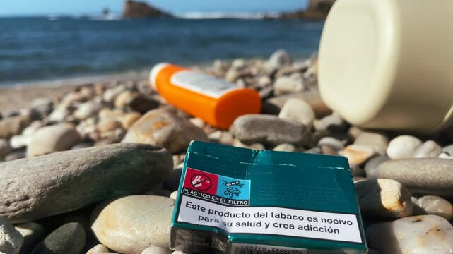 Discarded cigarette box and plastic waste on rocky beach shore, symbol of pollution